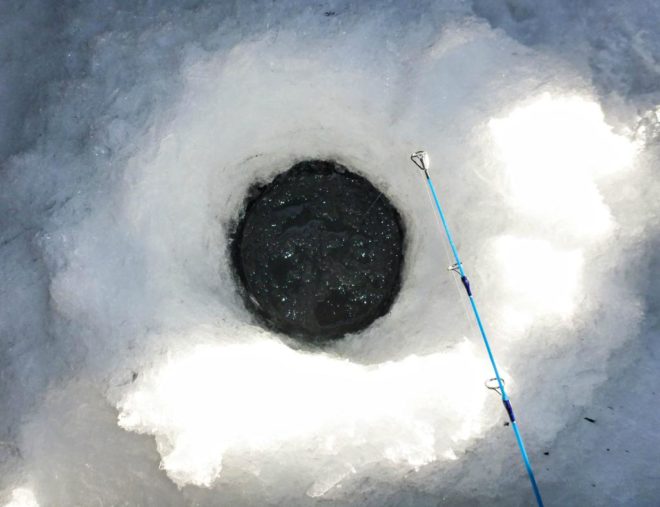 A blue fishing rod sits overtop of an ice fishing hole.