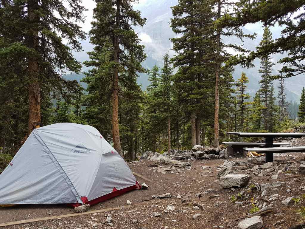 A tent sits on a dirt pad with trees and mountains in the background