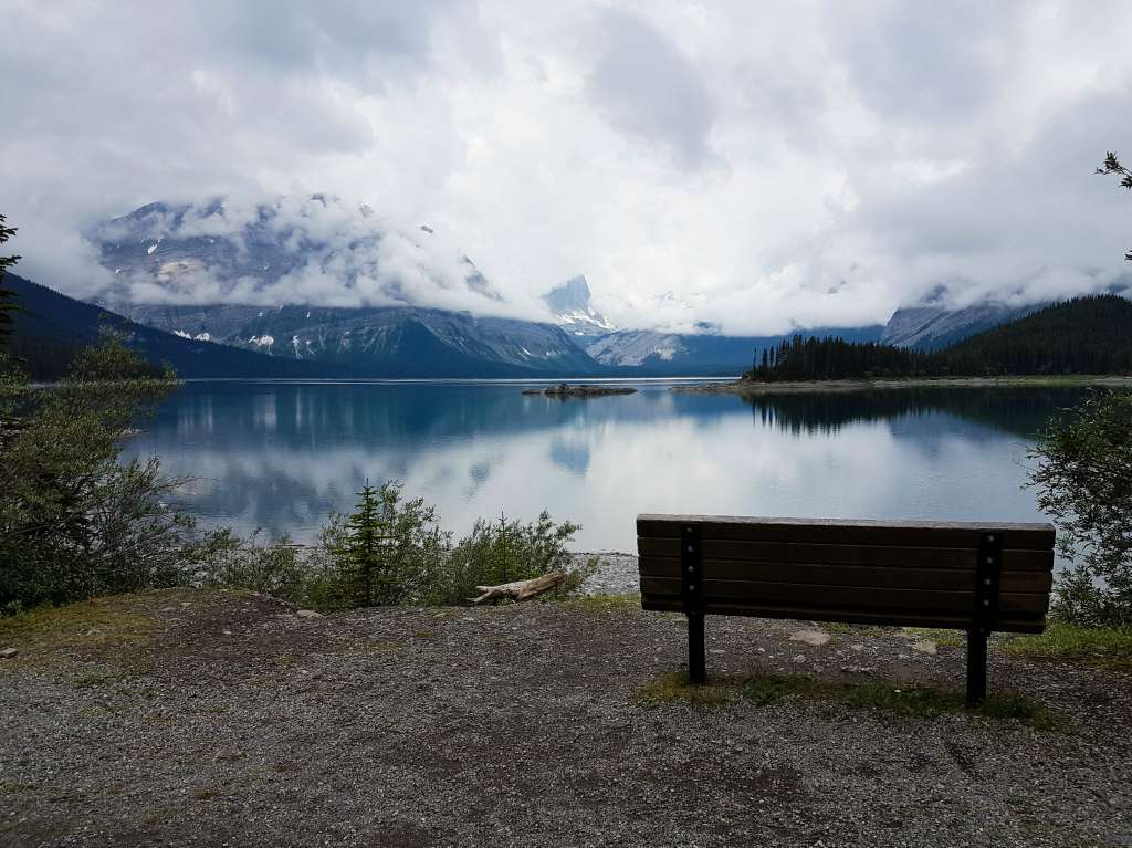 A bench sits on a lake shore with mountains and clouds in the background