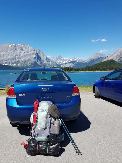 A backpack leans up against a car in a parking lot backed by mountains