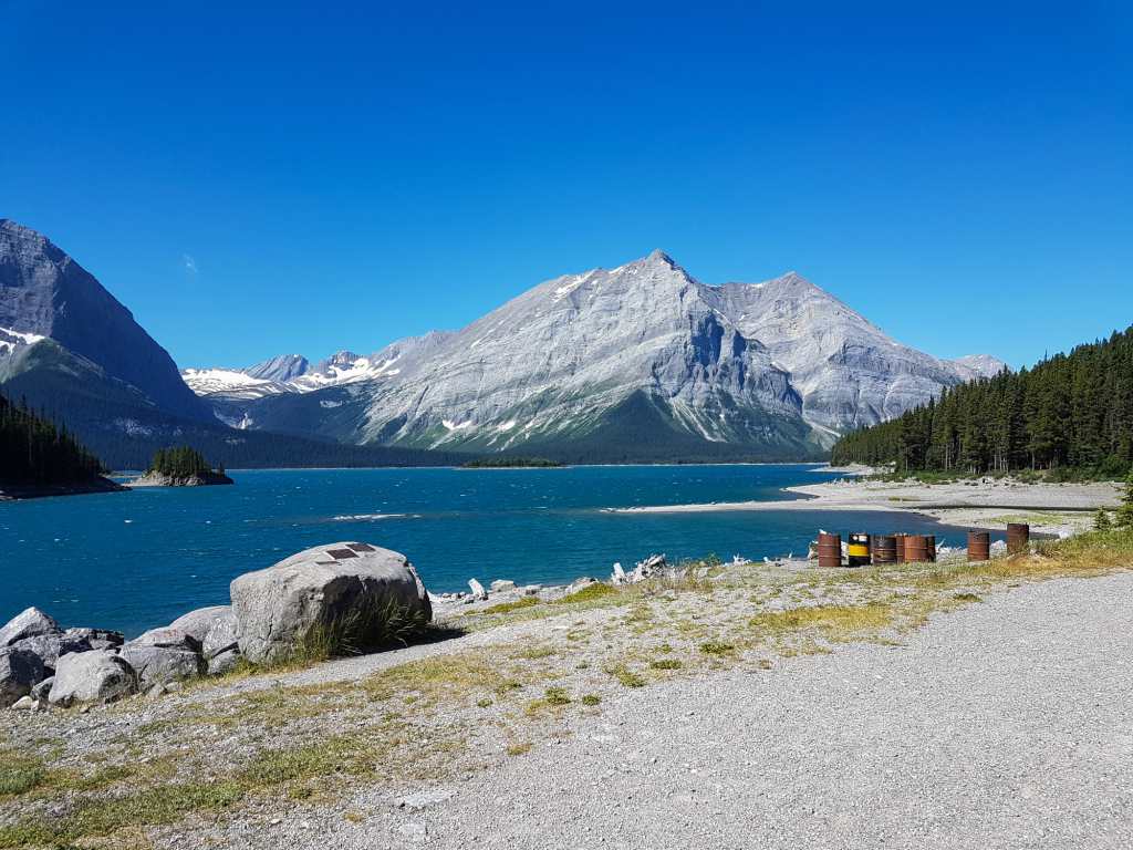 Barrels sit on the shoreline by a lake with mountains in the background