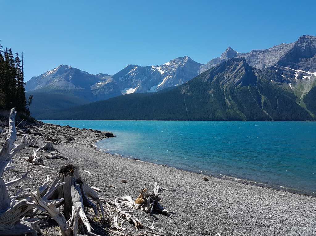 A rocky beach lines a lake with mountains in the background