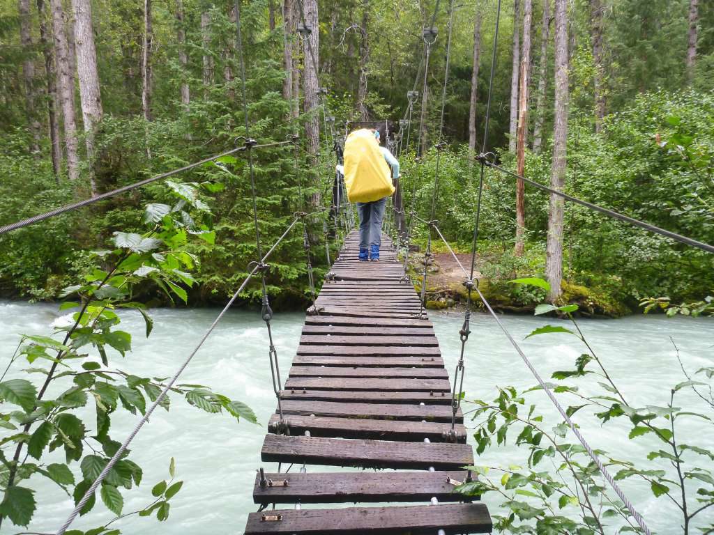 A woman with a large yellow backpack crosses a suspension bridge over the river