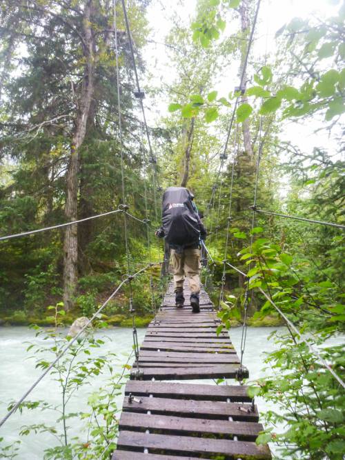 A man with a backpack crosses a suspension bridge over a river
