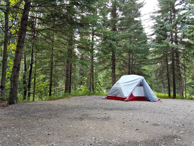 A red and grey tent is set up on a gravel pad surrounded by trees.