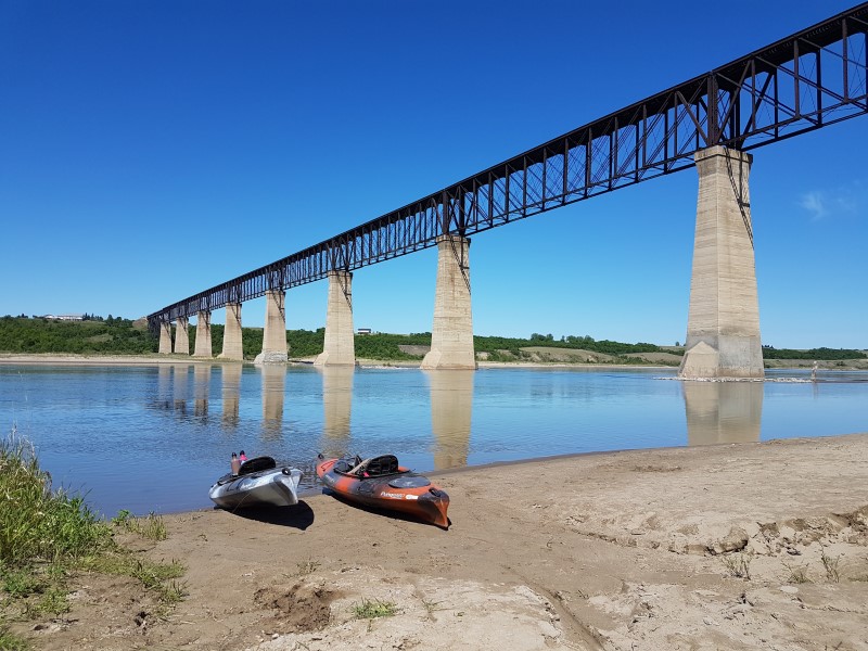Two kayaks sit on the riverbank under a bridge on the Chief Whitecap Waterway.