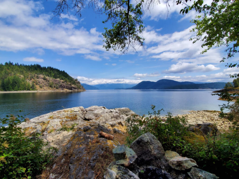 Blue skies and white clouds overhang a bright blue ocean bay. Low mountains appear in the background while a rocky shore lines the foreground.