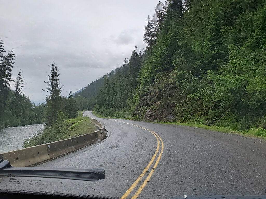 The view out a windshield shows the Sea to Sky Highway snaking between a river on the left and a steep tree covered mountain on the right.