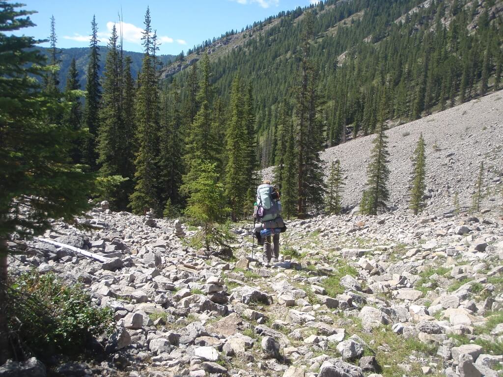 A backpacker walks along a rocky trail with trees and mountains in the background 