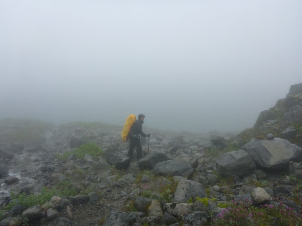A backpacker moves along a rocky trail while fog and rain obscure the view behind her. 