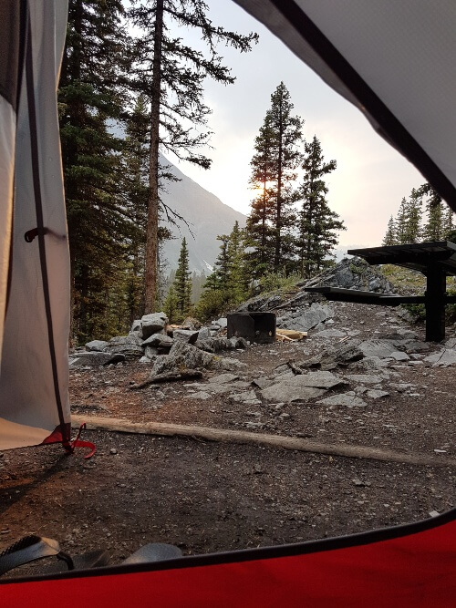 A view from a tent door shows a fire ring and picnic table with a mountain and trees in the background at sunset