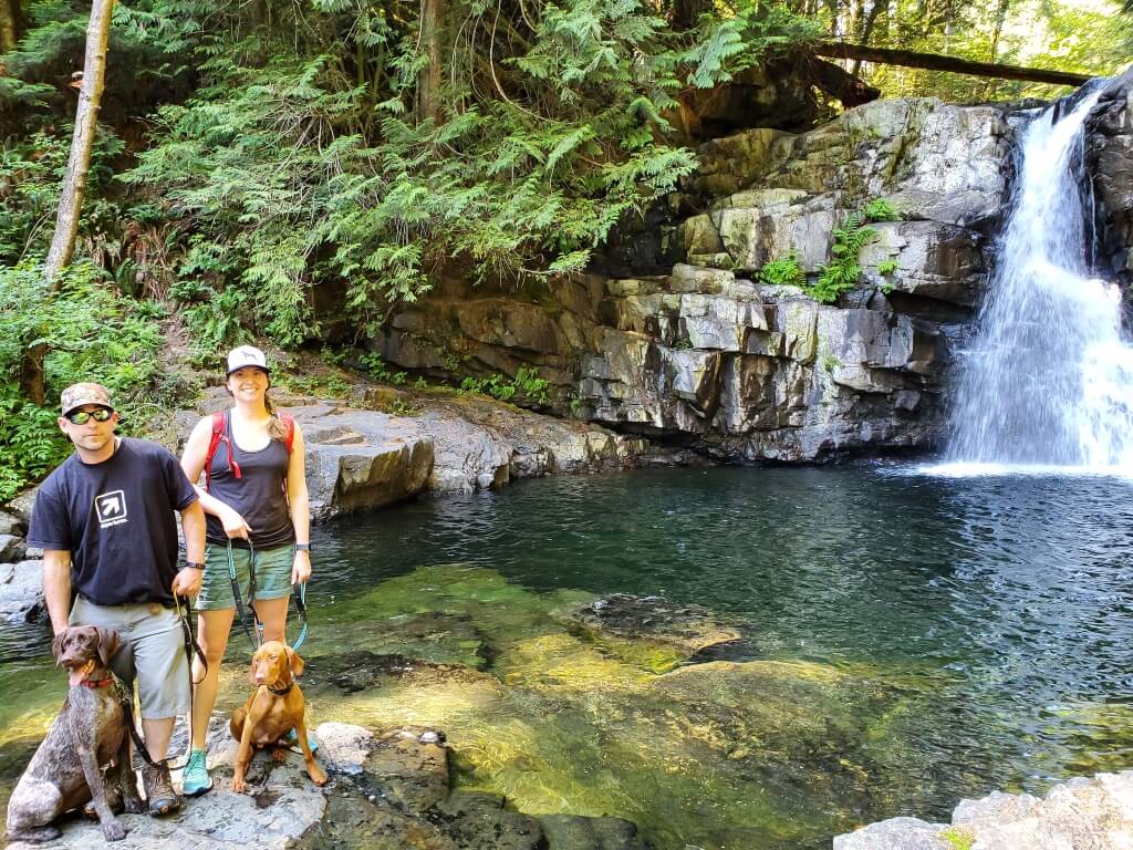 Mark and Laura stand on a rock holding two dogs on leashes with a waterfall and forested hillside in the background