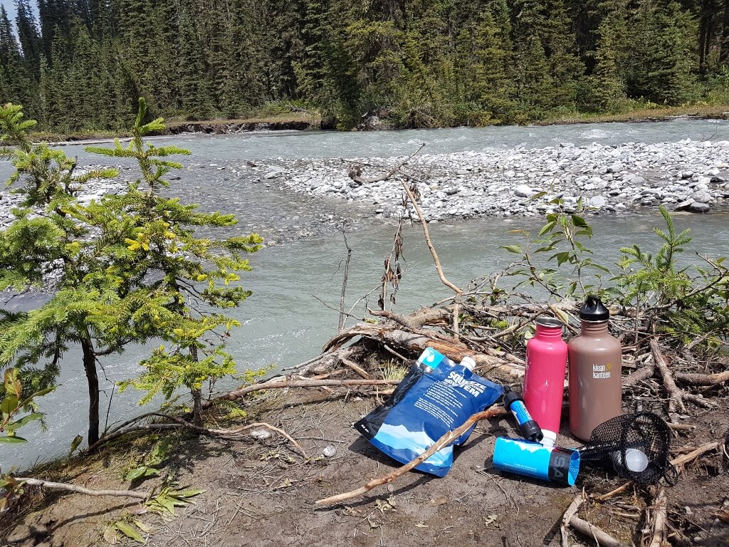 A water filter and two water bottles sit on the ground beside a river