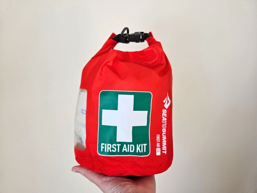 A bright red first aid kit with a green and white first aid symbol is held in the palm of a hand.