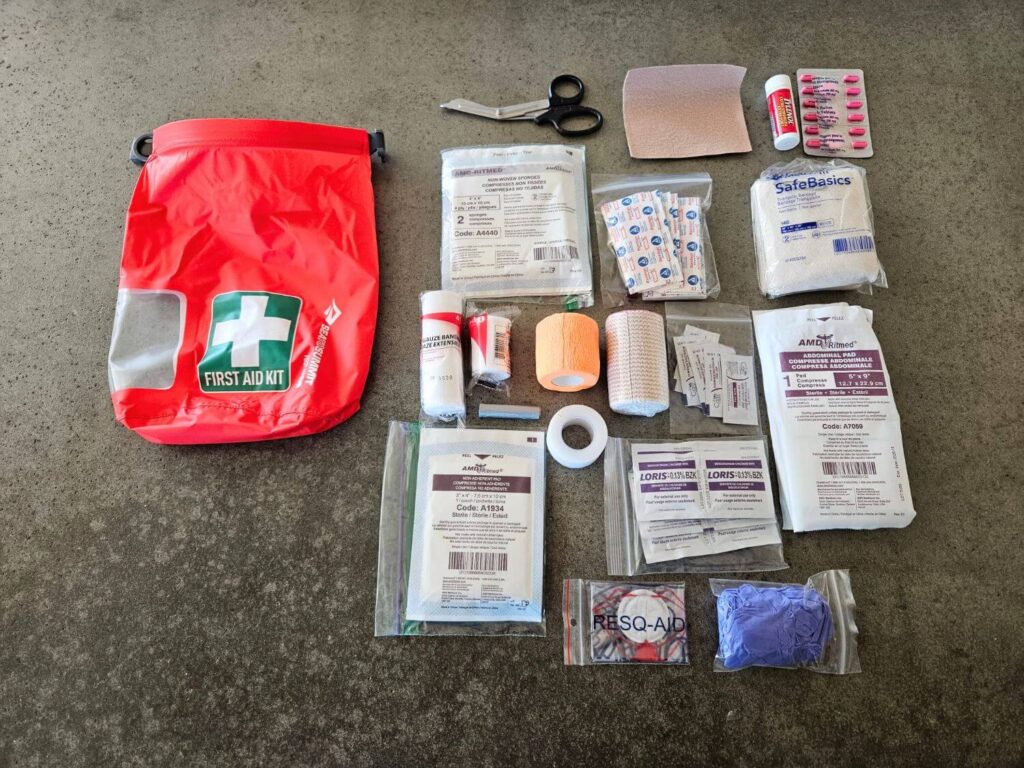 The contents of a first aid kit sit on a counter beside a red, green and white dry bag.