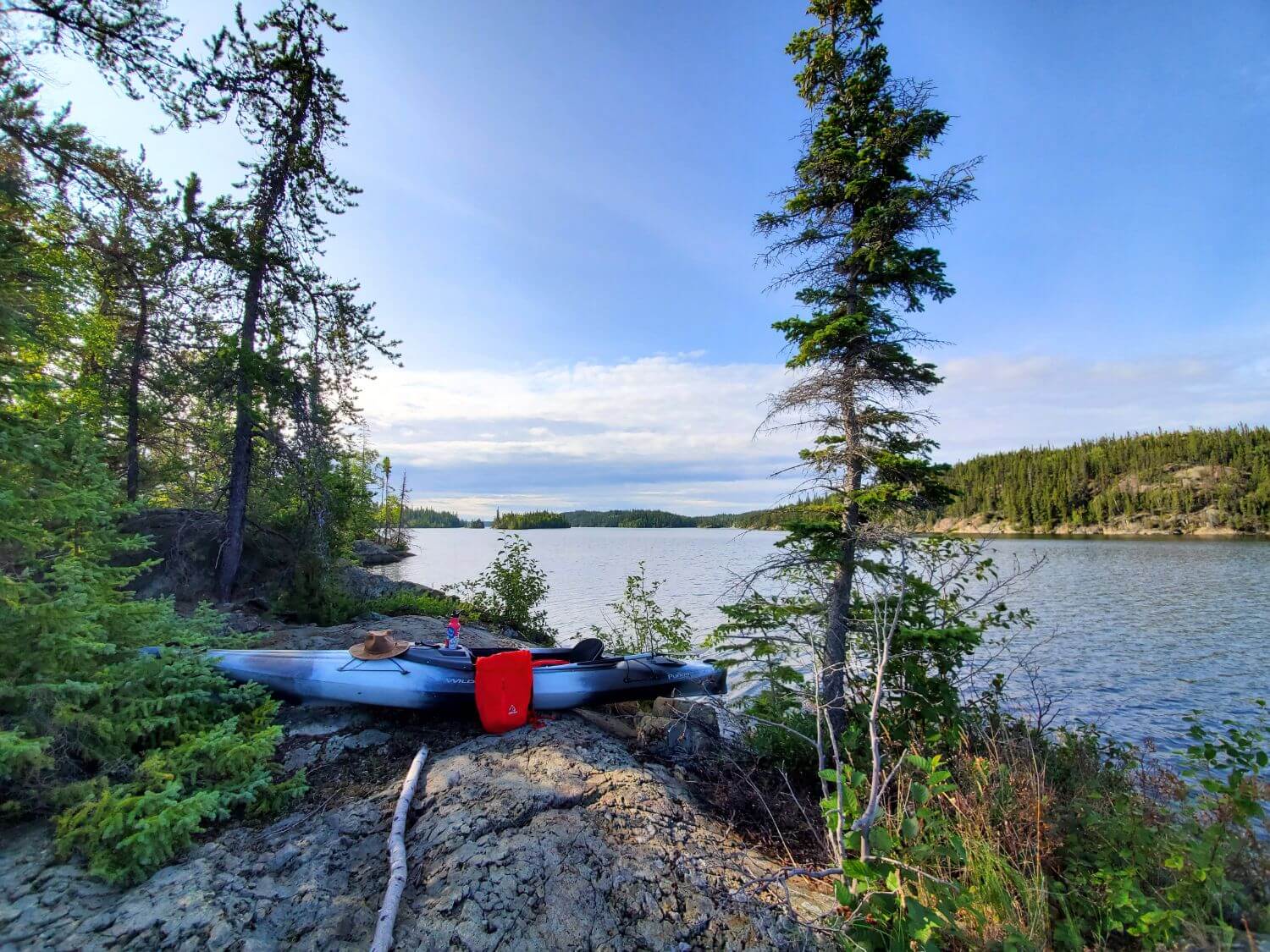 A grey kayak sits on a rocky shoreline surrounded by spruce trees. A hat, water bottle, and red backpack sit atop the kayak. A blue sky and lake are visible in the background.